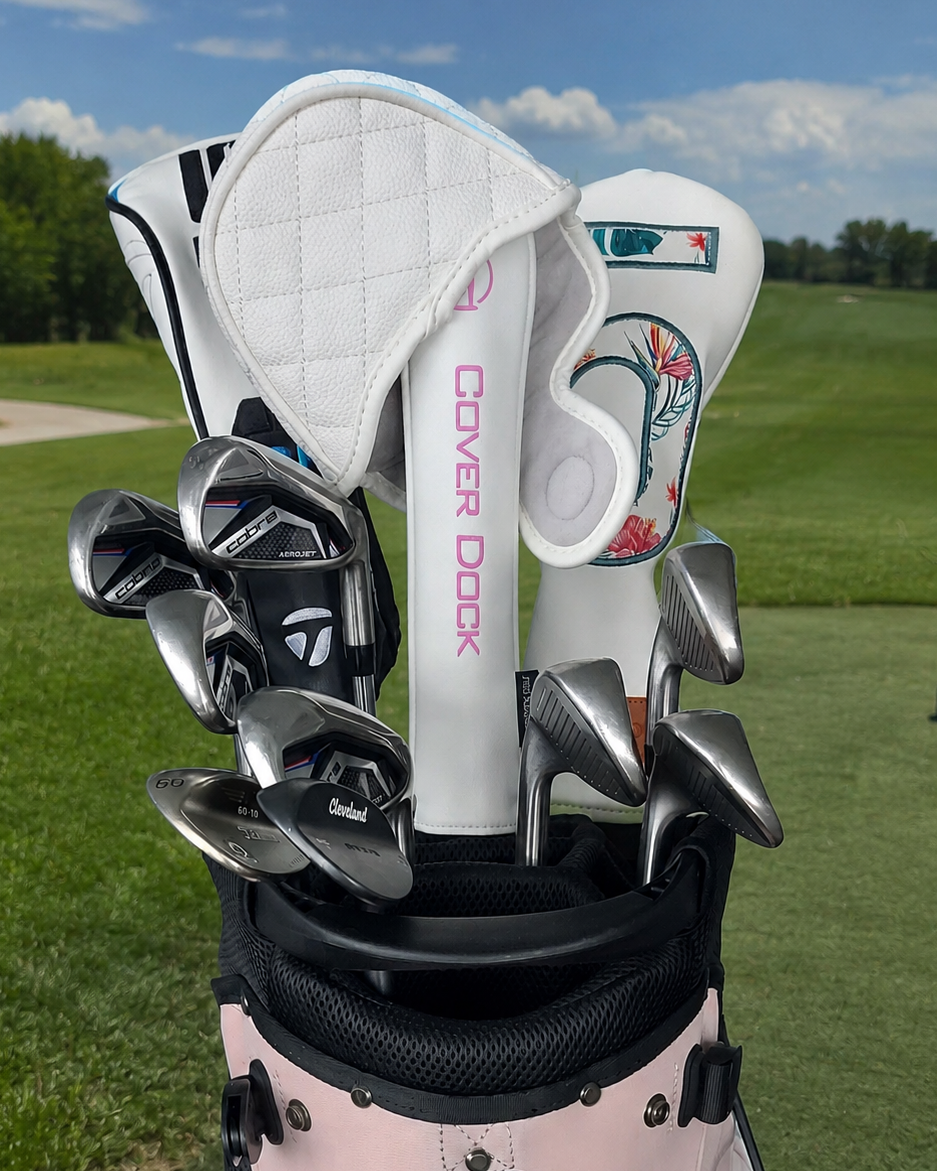 A golf bag on a golf course with several golf clubs sticking out. The bag is light pink and black, and the club heads are visible, including a white headcover with pink 'COVER DOCK' text and another white headcover with floral artwork.