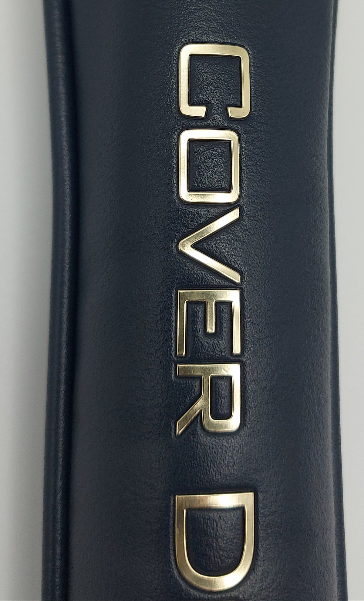 Close-up of a dark navy blue textured leather or faux-leather cylindrical item with large metallic gold 'COVERD' lettering printed vertically on the side.