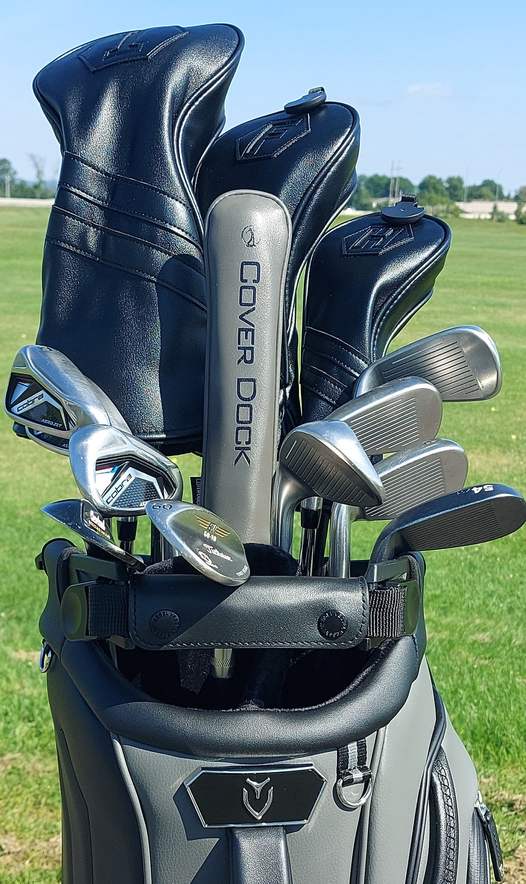 A golf bag on a grassy golf course with several golf clubs and headcovers inserted. The bag is dark gray and black, and one of the headcovers reads COVER DOCK.