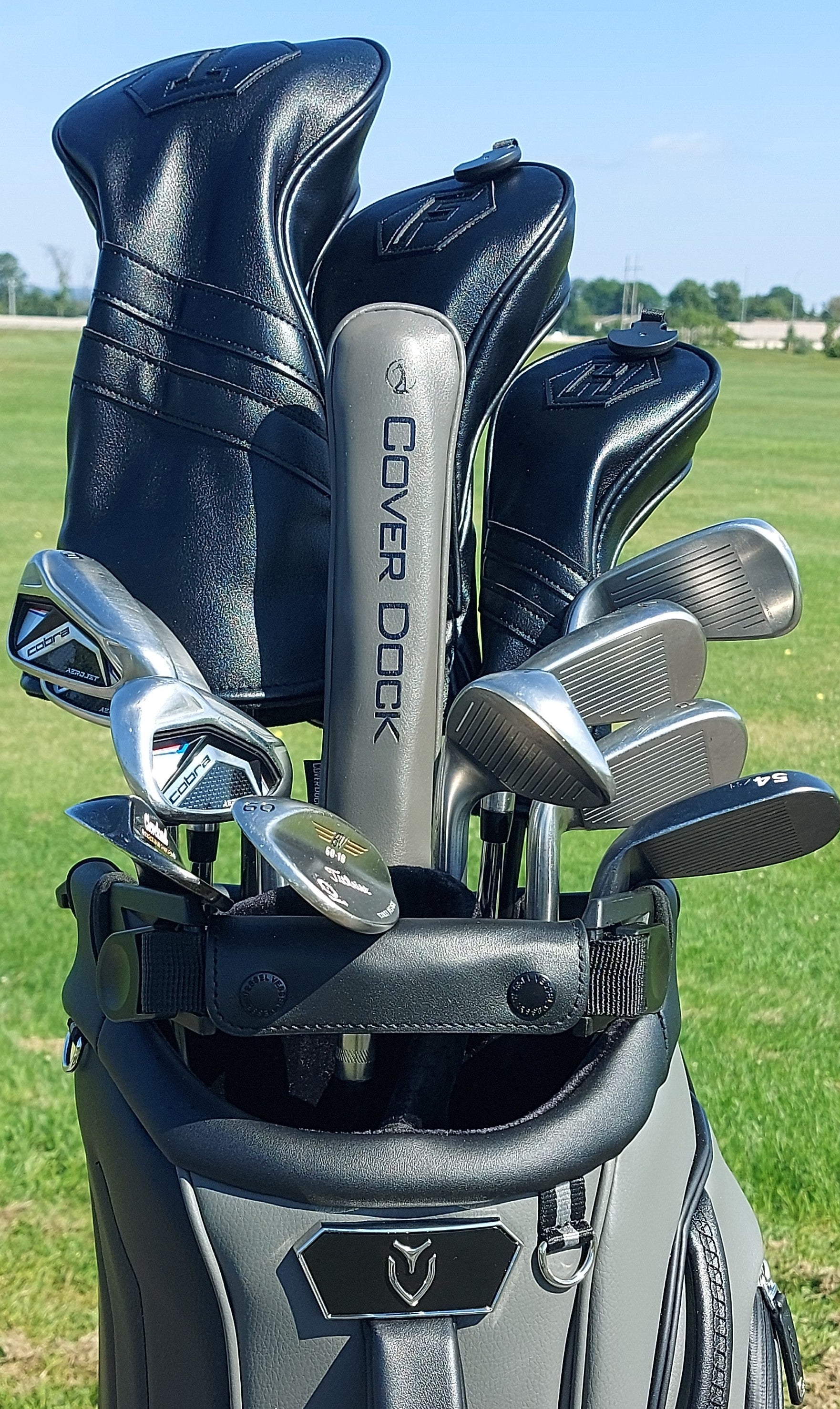 A golf bag on a grassy golf course with several golf clubs and headcovers inserted. The bag is dark gray and black, and one of the headcovers reads COVER DOCK.