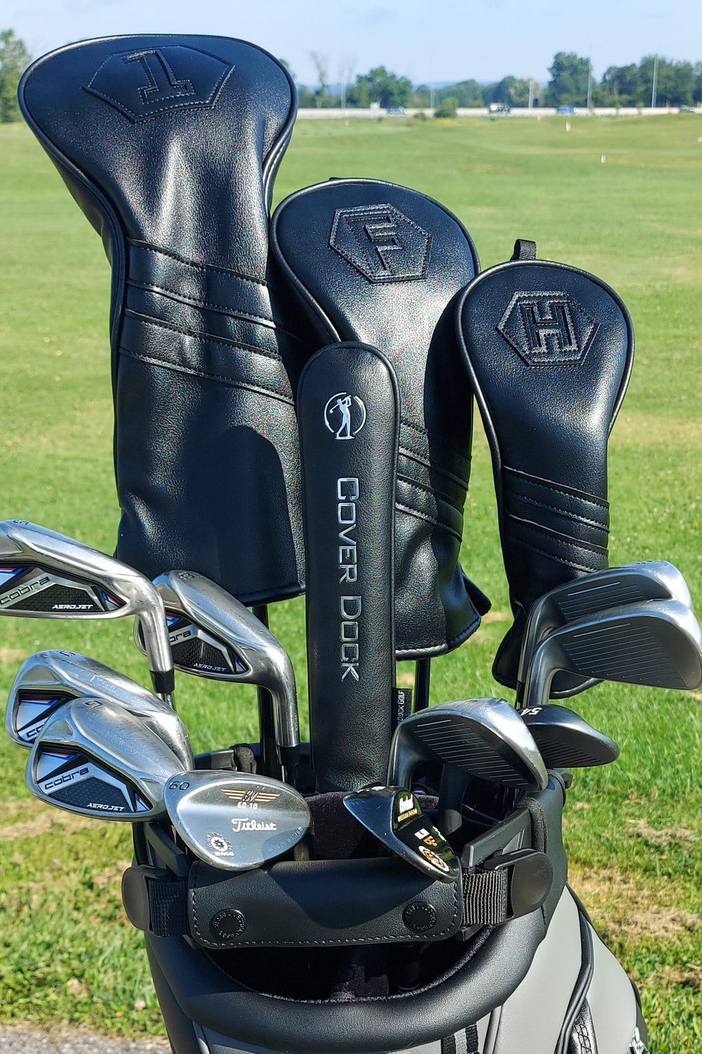 A golf club set in a black stand bag on a grassy golf course. The set includes black leather club headcovers with embossed logos, a black cover dock, and several silver metal golf clubs.