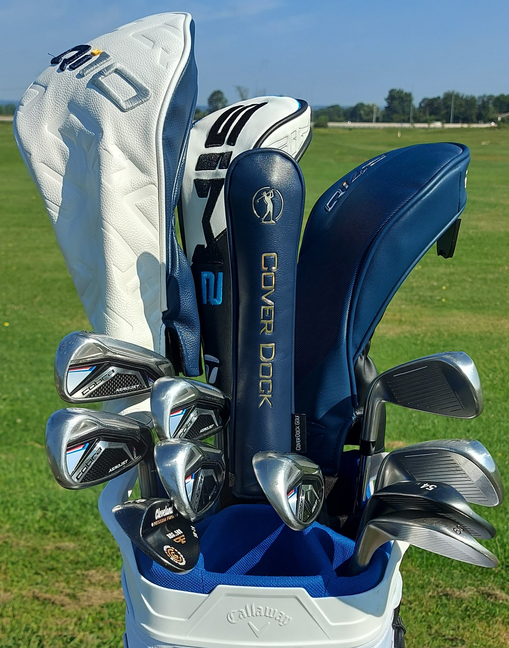 A Callaway golf bag on a grassy golf course holding several golf clubs with blue and white Callaway cover heads, including an iron and a wood.