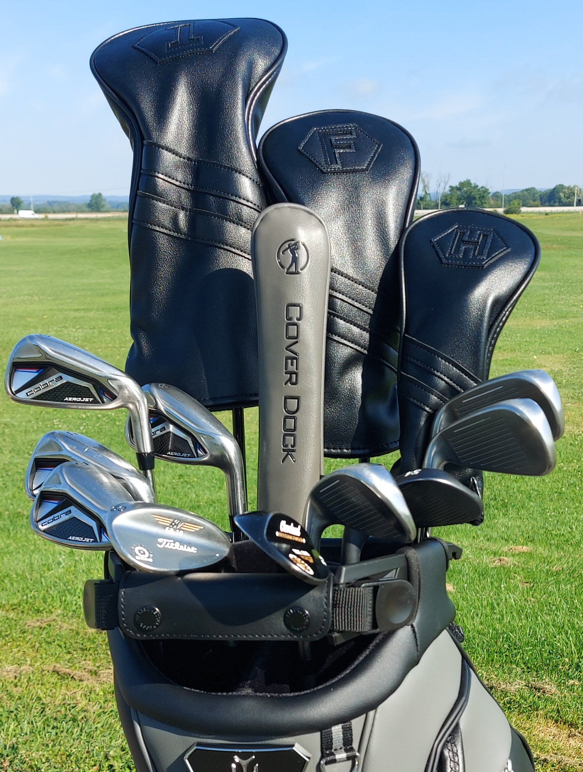 A golf bag on a grassy golf course with several golf clubs and black leather headcover sleeves standing upright. Visible branding includes Fidelity on the club shaft and Cover Dock on the bag.