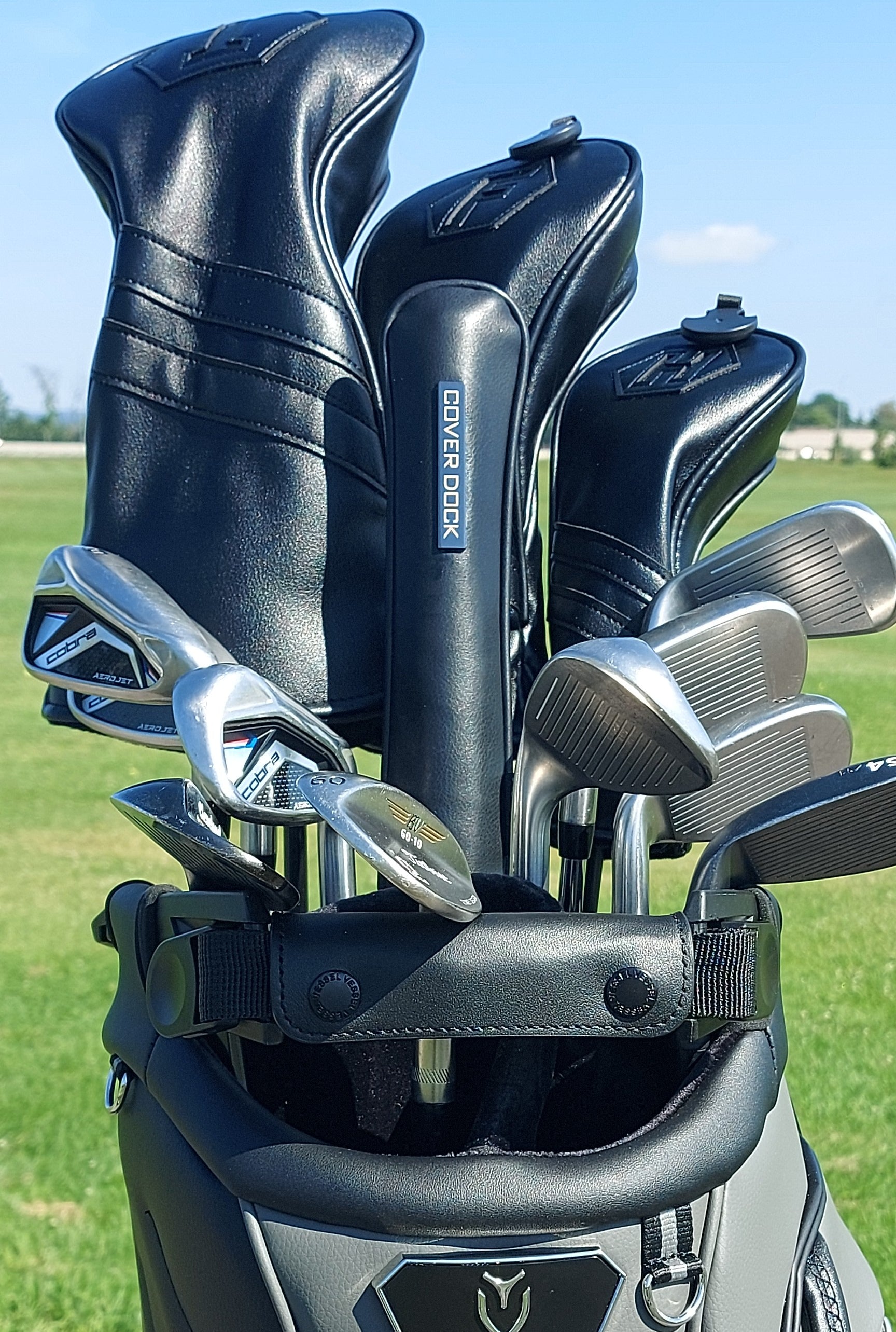 A golf bag filled with golf clubs, including  irons and  headcover and several black and silver club heads, photographed outdoors on a grassy golf course under a blue sky. metallic badge with cover dock written on it inserted in the bag to hold golf club head covers
