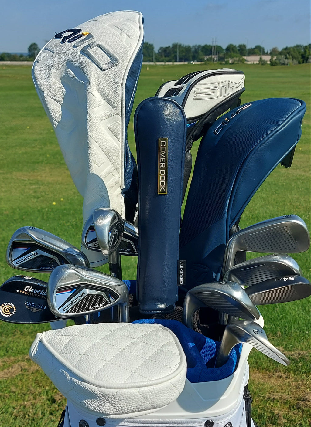 A golf club set in a white and blue golf bag on a grassy golf course. The set includes a white  headcover, a navy blue neck cover with a gold 'Callaway' logo, and several silver metal clubs.