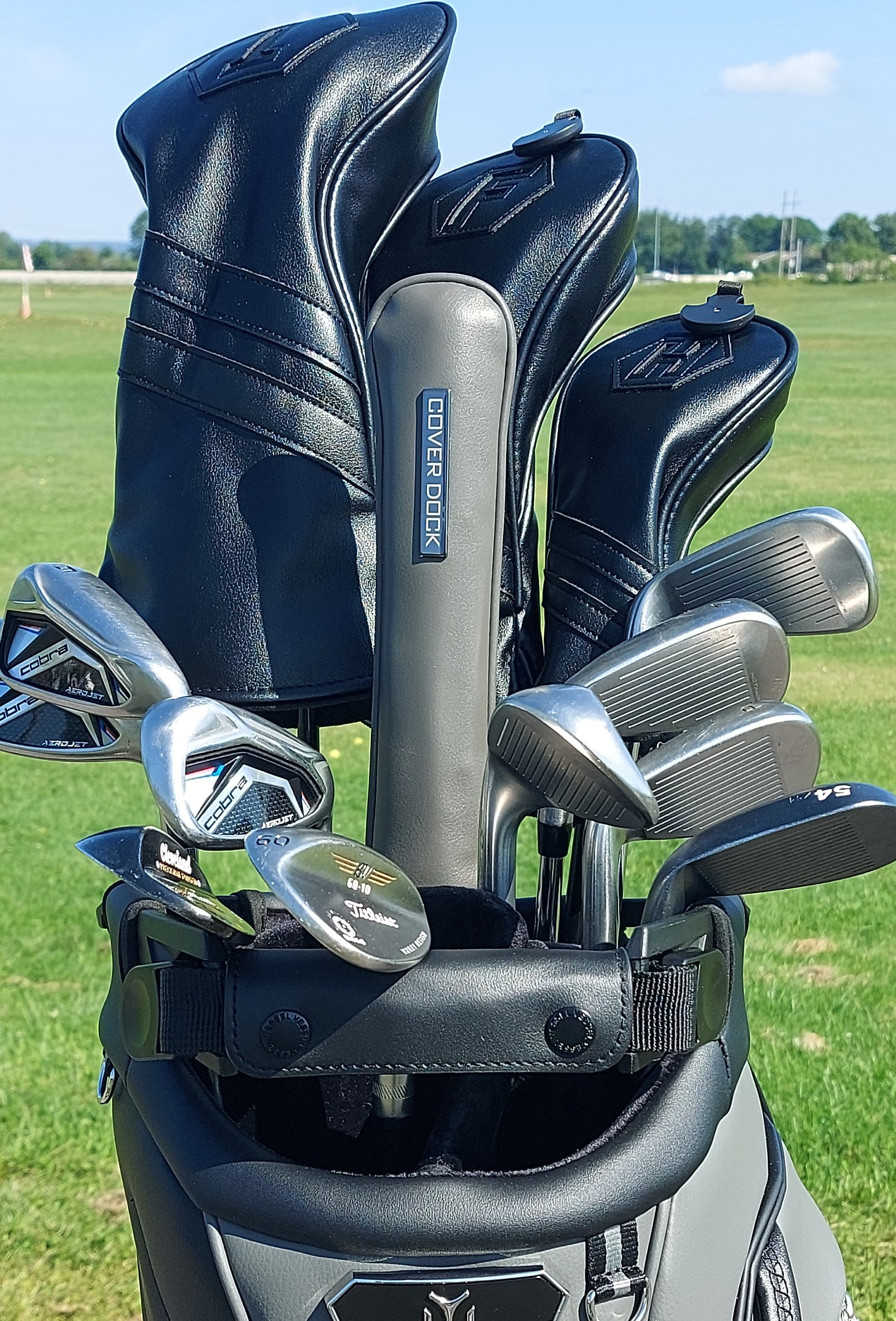 A golf club bag filled with golf clubs on a grassy golf course. The bag has black leather headcovers, and the clubs include silver metal irons and woods.