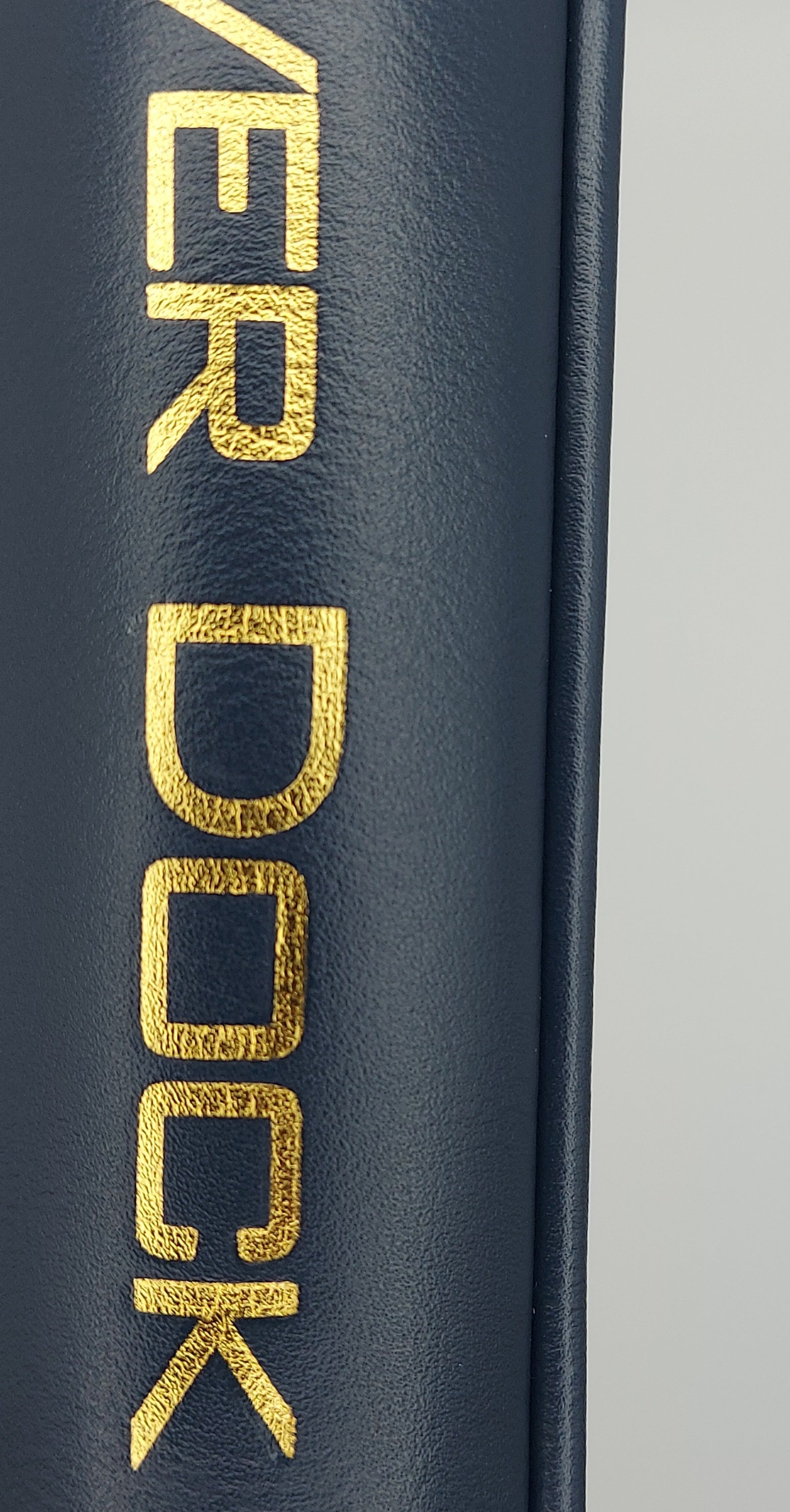 Close-up of a dark navy blue textured surface with large metallic gold letters spelling part of a brand name vertically, against a light gray background.