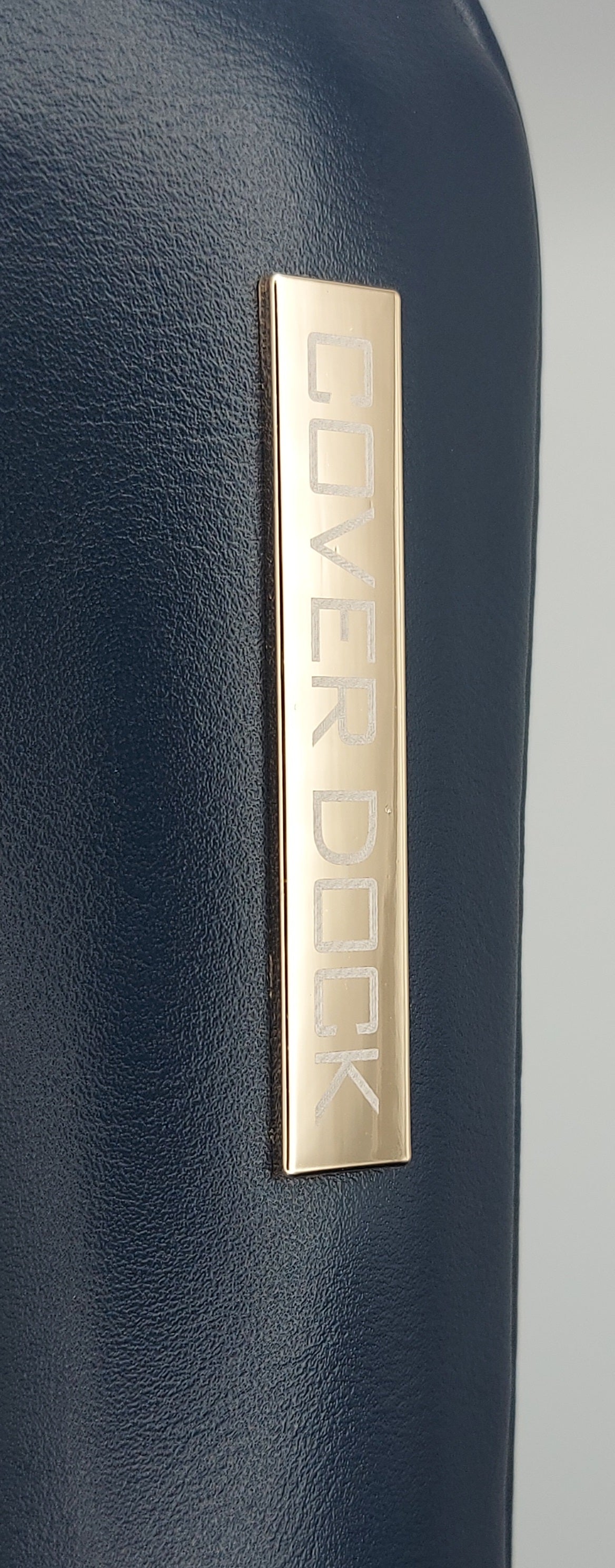 Close-up of a dark navy blue textured surface with a vertical gold metal plaque engraved with the brand name COVER DOCK.