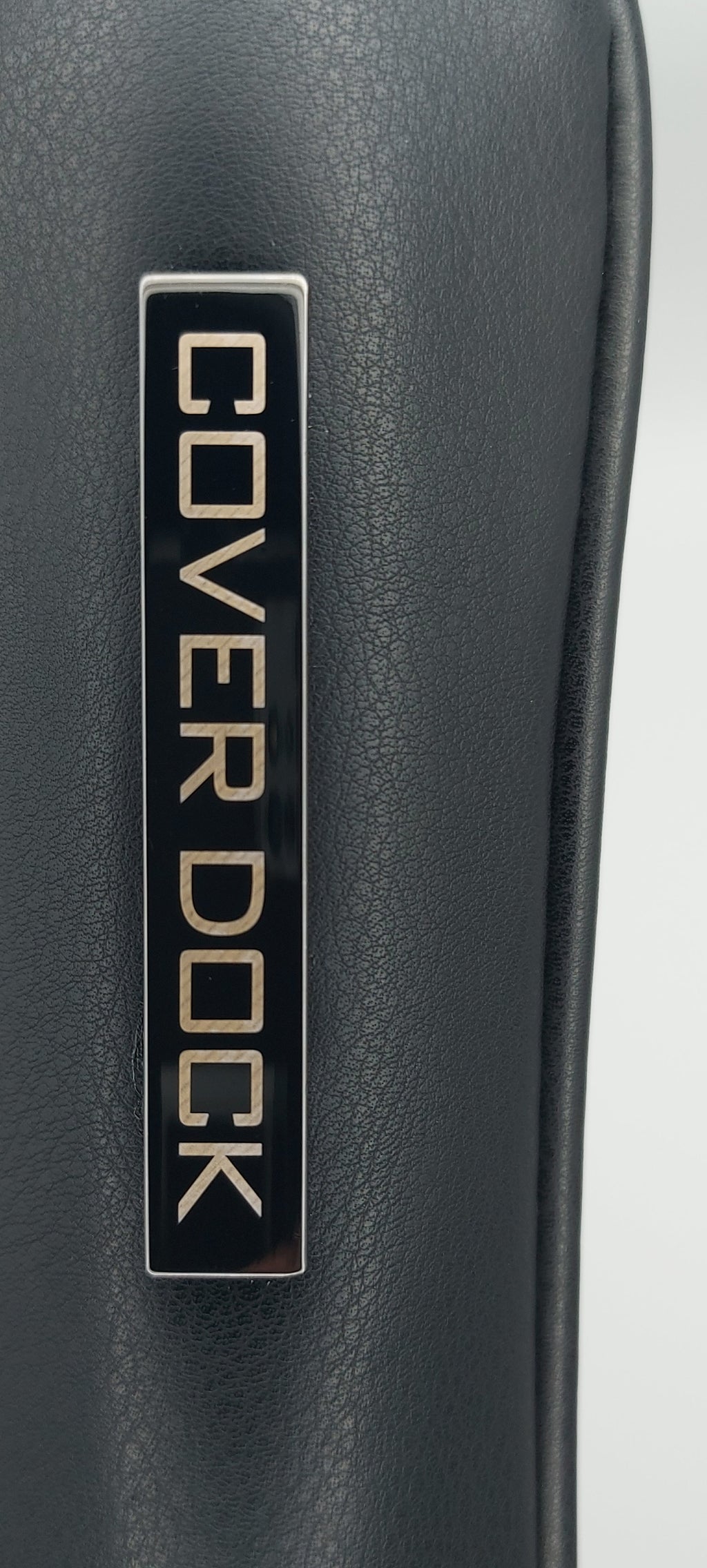 Close-up of a black leather or vegan leather surface with a rectangular metal plaque reading COVER DOCK in gold lettering.