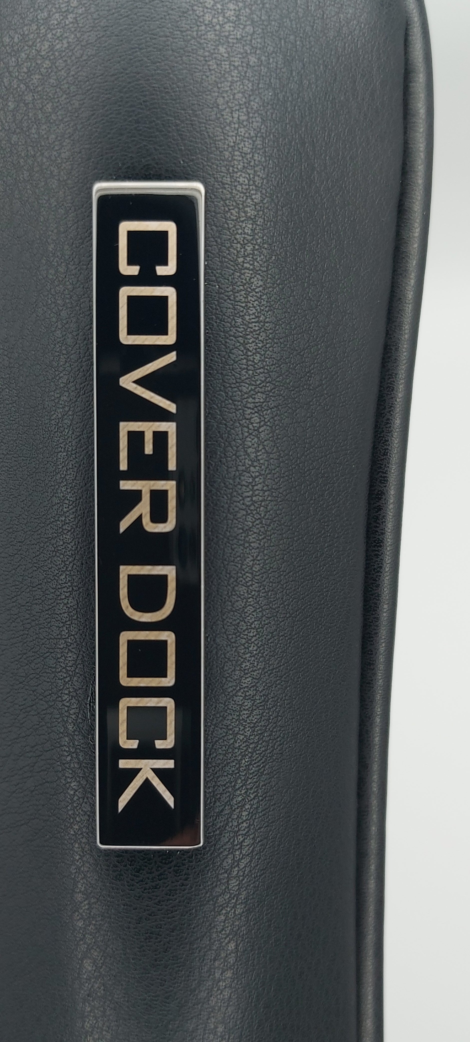 Close-up of a black leather or vegan leather surface with a rectangular metal plaque reading COVER DOCK in gold lettering.